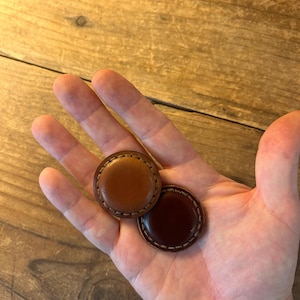 May include: Two round leather buttons, one brown and one dark brown, with stitching around the edges.
