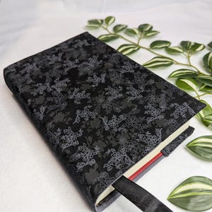 May include: A black book cover with a floral pattern in shades of gray. The cover has a black ribbon bookmark. The book is resting on a white surface with green and white leaves.
