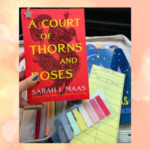 May include: A bright red book titled A Court of Thorns and Roses by Sarah J. Maas with a star-shaped metal bookmark, a yellow annotation key card, and a set of colorful adhesive page markers in various pastel and vibrant shades.