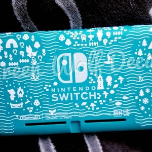May include: Teal Nintendo Switch Lite console with a white illustrated design. The design includes various icons such as trees, houses, and people. The Nintendo Switch logo is visible.