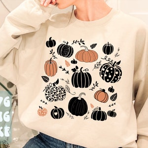 May include: Beige sweatshirt with a pumpkin design. The design features various illustrated pumpkins in black, orange, and white, with leaf and vine accents. The sweatshirt also has the text "SVG PNG DXF" in a light blue color.