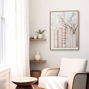 May include: A framed print of a white bookshelf with a row of antique books and a delicate branch of white blossoms. The print is hanging on a white wall in a room with a white armchair and a wooden side table.