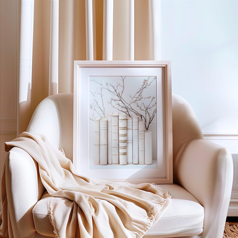 May include: A framed print of a still life featuring a stack of white books with brown spines and a delicate branch of dried flowers. The print is displayed on a white armchair with a beige throw blanket.