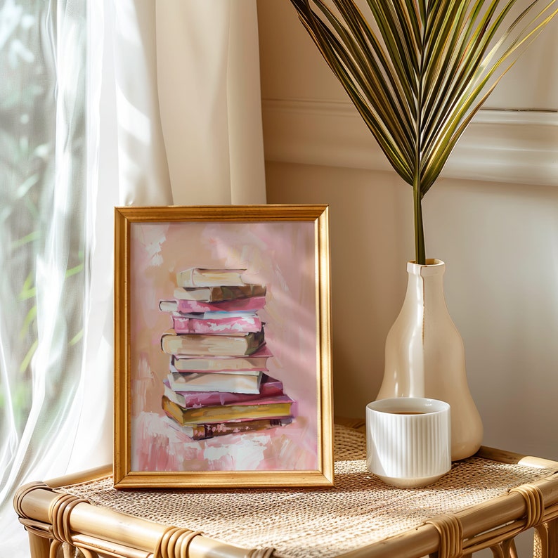 May include: A framed painting of a stack of books with pink, brown, and white covers. The books are arranged in a pile on a pink background. The painting is in a gold frame and is sitting on a table with a vase and a cup.