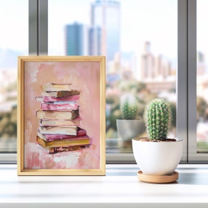 May include: A framed painting of a stack of books in shades of pink and brown on a peach background. The painting is on a white surface with a cactus in a white pot.