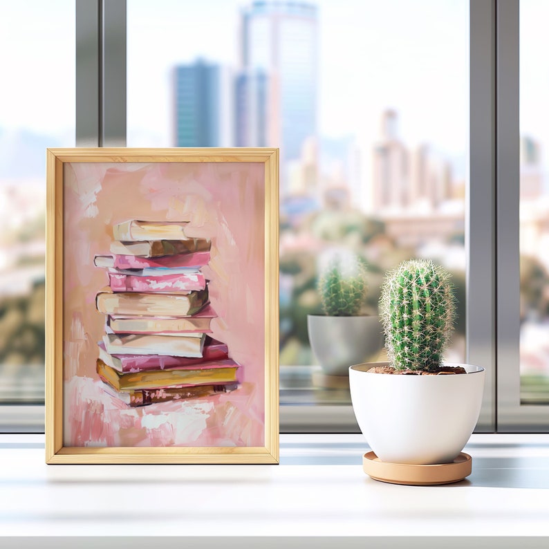 May include: A framed painting of a stack of books in shades of pink and brown on a peach background. The painting is on a white surface with a cactus in a white pot.