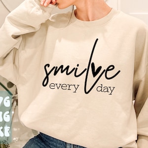 Puede incluir: Una sudadera beige con el texto "smile every day" en letra negra con un pequeño corazón en el medio de la palabra "smile".