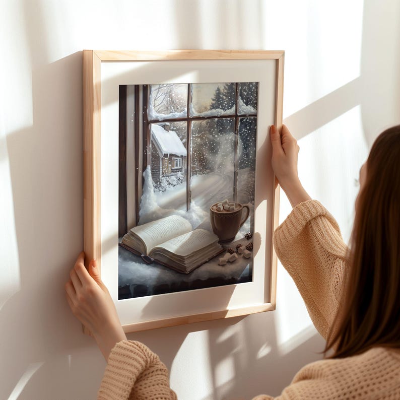 May include: A framed print of a winter scene with a cozy cottage, a book, and a mug of hot chocolate. The print is hanging on a white wall and a person's hand is reaching out to touch it.