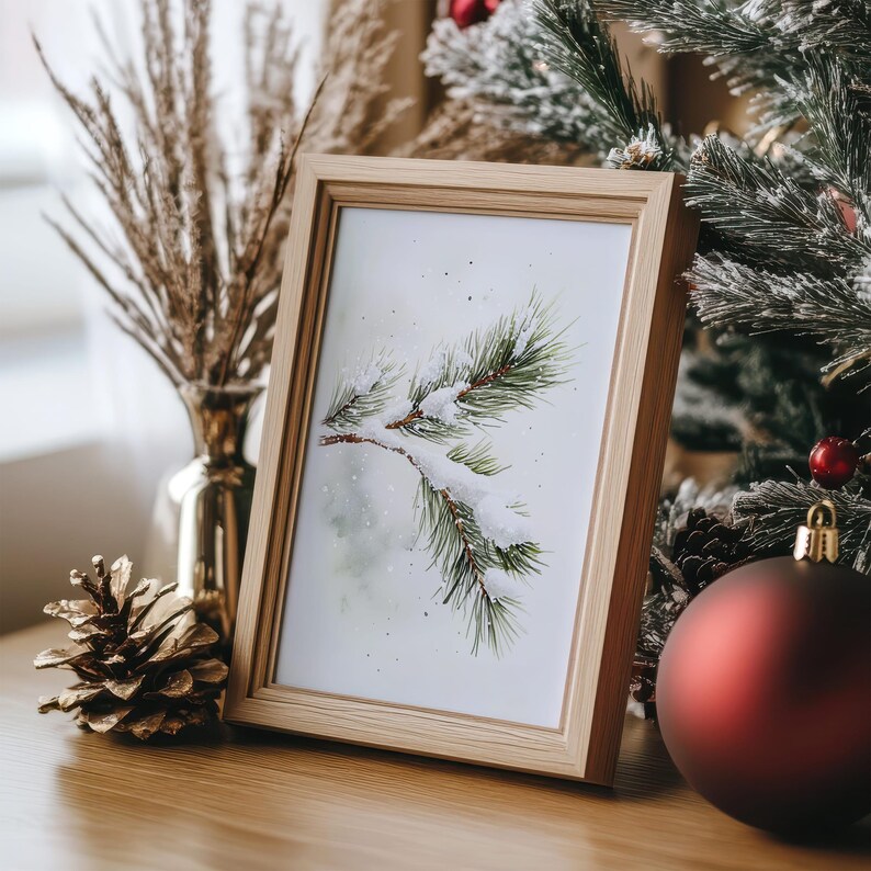 May include: A framed watercolor painting of a snow-covered pine branch with a white background. The frame is light brown wood.