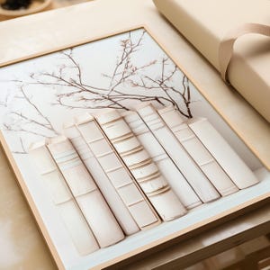May include: A framed print of a row of white books with a delicate floral design in the background. The books are arranged in a stack and have a vintage, antique look.