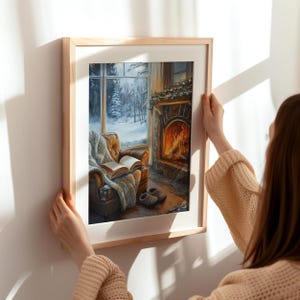 Peut inclure: Une peinture encadr&eacute;e repr&eacute;sentant un salon confortable avec une chemin&eacute;e, une fen&ecirc;tre avec de la neige &agrave; l'ext&eacute;rieur et un fauteuil confortable avec un livre dessus. La peinture est dans un cadre en bois et est accroch&eacute;e &agrave; un mur blanc.