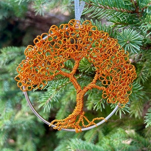 May include: An orange wire tree ornament with a silver metal ring and a light blue ribbon. The tree design is detailed and intricate, set against a backdrop of green pine needles. The ornament is suitable for a Christmas tree.