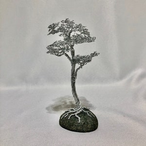 May include: A sculpture of a tree crafted from silver-colored wire, featuring a twisted trunk and intricate wire loops forming the canopy. The tree is anchored to a smooth, dark gray, rounded stone base that displays visible root-like wire details.