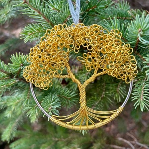 May include: A gold-colored wire tree ornament with a silver-colored metal ring and a light blue ribbon for hanging. The tree design features a detailed trunk and branches with a circular crown. The ornament is displayed against a backdrop of green pine needles.