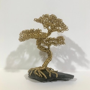 May include: A gold wire sculpture of a bonsai tree with a twisted trunk and intricate branches. The tree is sitting on a black stone base.