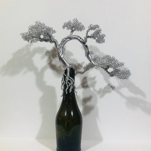 May include: A sculpture of a bonsai tree crafted from silver-colored wire, featuring a twisted trunk and intricate foliage clusters. The wire roots extend downward from the base of the tree, wrapping around the neck of a dark green glass bottle.