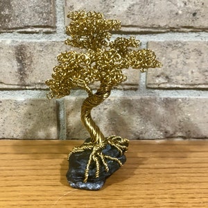 May include: A gold wire sculpture of a bonsai tree with a twisted trunk and branches. The tree is sitting on a dark gray rock.