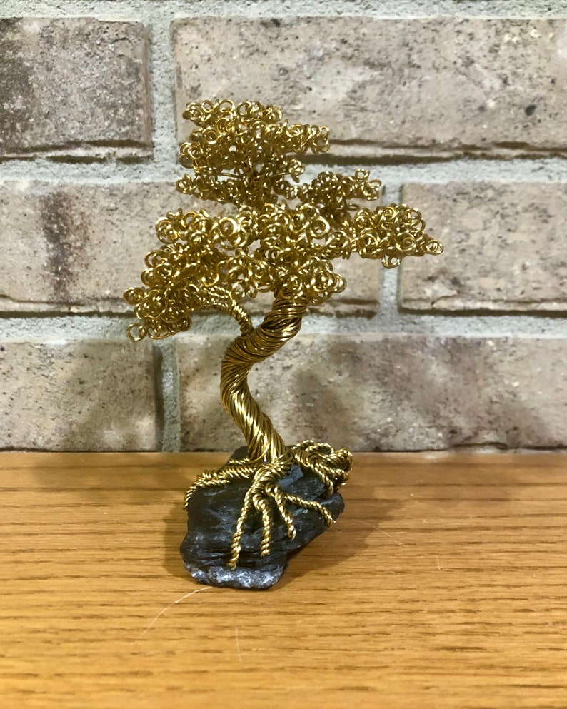 May include: A gold wire sculpture of a bonsai tree with a twisted trunk and branches. The tree is sitting on a dark gray rock.
