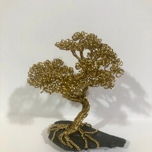 May include: A gold wire sculpture of a tree with a twisted trunk and branches. The tree is sitting on a dark gray rock base.