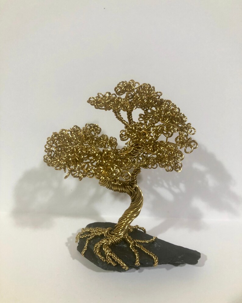 May include: A gold wire sculpture of a tree with a twisted trunk and branches. The tree is sitting on a dark gray rock base.