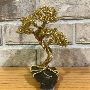 May include: A gold wire sculpture of a bonsai tree with a twisted trunk and branches. The tree is sitting on a black rock base.