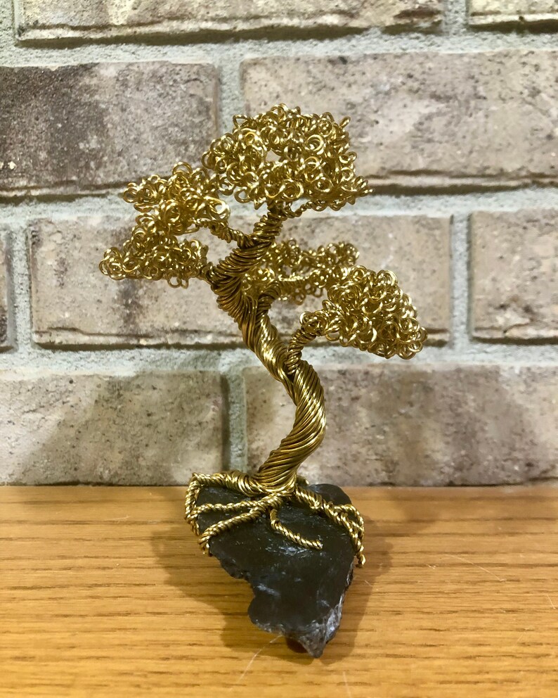 May include: A gold wire sculpture of a bonsai tree with a twisted trunk and branches. The tree is sitting on a black rock base.