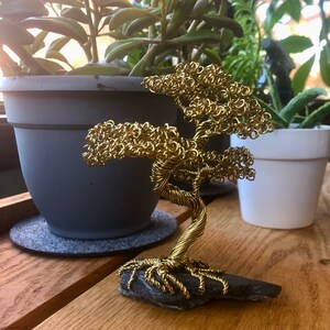 May include: A gold wire sculpture of a bonsai tree on a black slate base. The tree is made of tightly wound wire and has a realistic shape.