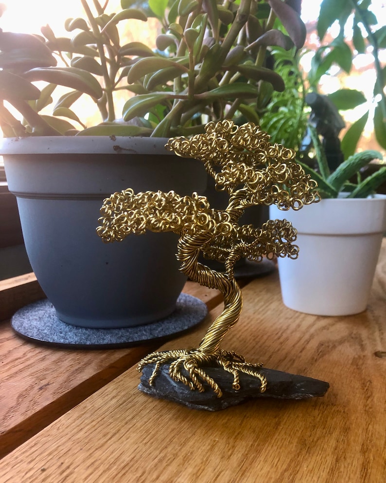 May include: A gold wire sculpture of a bonsai tree on a black slate base. The tree is made of tightly wound wire and has a realistic shape.