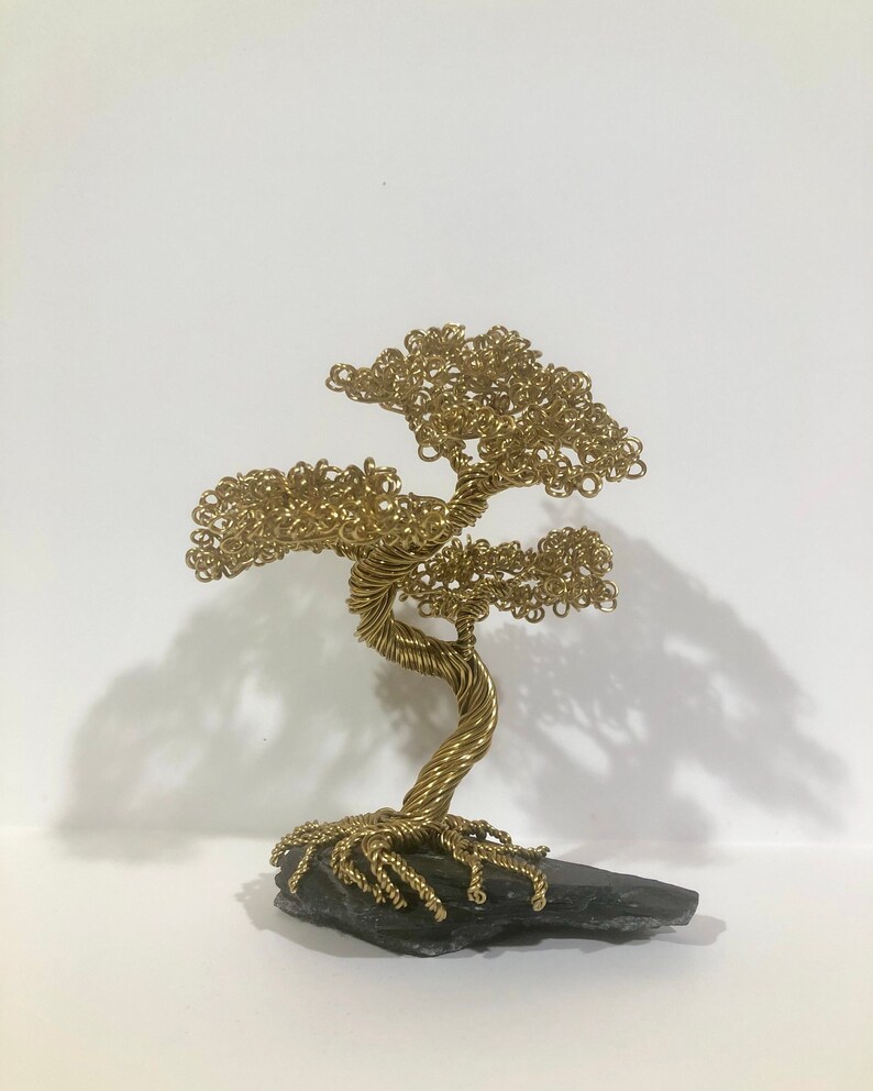 May include: A gold wire sculpture of a bonsai tree with a twisted trunk and intricate branches. The tree is sitting on a black stone base.