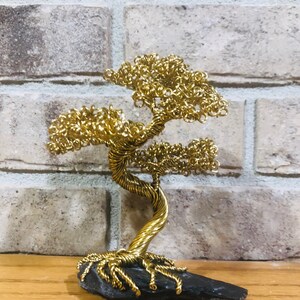 May include: A gold wire sculpture of a bonsai tree with a twisted trunk and branches. The tree is sitting on a dark gray rock base.