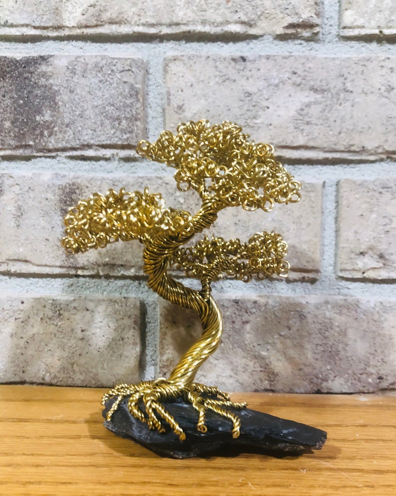 May include: A gold wire sculpture of a bonsai tree with a twisted trunk and branches. The tree is sitting on a dark gray rock base.