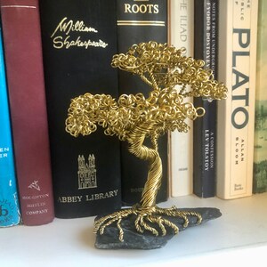 May include: A gold wire sculpture of a tree with a black slate base. The sculpture is sitting on a shelf with a row of books behind it.