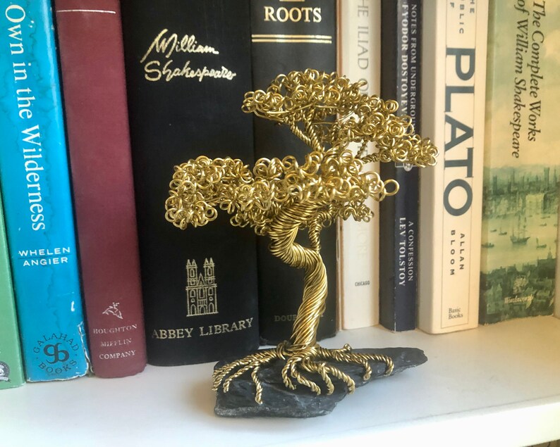 May include: A gold wire sculpture of a tree with a black slate base. The sculpture is sitting on a shelf with a row of books behind it.