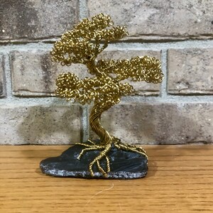 May include: A gold wire sculpture of a bonsai tree on a black slate base. The tree has a twisted trunk and branches with curled leaves.