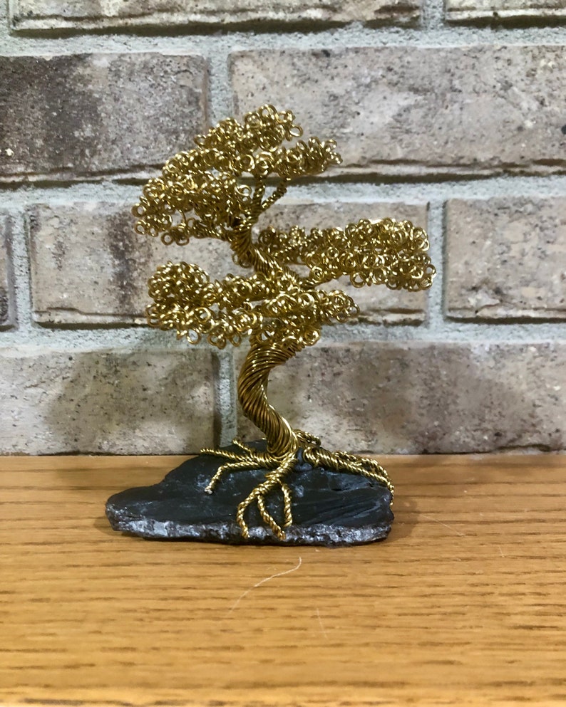 May include: A gold wire sculpture of a bonsai tree on a black slate base. The tree has a twisted trunk and branches with curled leaves.