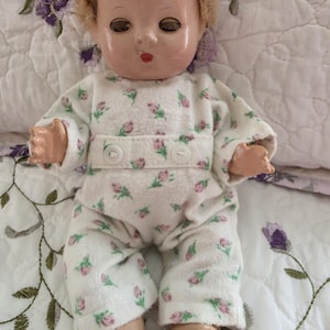 May include: A vintage doll with painted facial features, including closed eyes and red lips. The doll wears a white onesie with a pink floral pattern and has blonde, curly hair. The doll's hands and feet are painted a flesh tone.