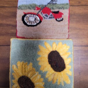 May include: Two handmade, square, textured rugs. The top rug features a red motorcycle on a tan background with a blue sky. The bottom rug displays two yellow sunflowers with brown centers on a green background.