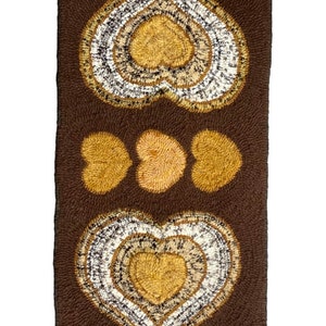 May include: A brown rug with a repeating pattern of three hearts. The hearts are made of a lighter brown and white yarn. The rug is approximately 24 inches wide and 48 inches long.
