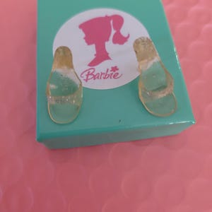 May include: A pair of miniature clear plastic sandals next to a teal box with a pink Barbie logo. The sandals have a thong-style design. The box is on a pink surface.