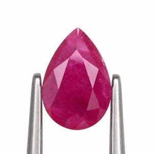 May include: A pear-shaped, faceted ruby gemstone with a deep, vibrant pinkish-red color. The gemstone is held securely between the two silver-toned metal prongs of a pair of tweezers, showcasing its clear, symmetrical cut and polished surface.