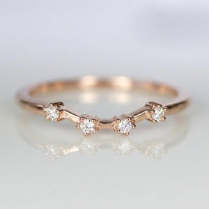Puede incluir: Anillo de banda de oro rosa con cinco peque&ntilde;as piedras preciosas claras. El anillo tiene un dise&ntilde;o curvo con las piedras preciosas dispuestas en una fila en la parte superior. El anillo se muestra sobre una superficie reflectante.