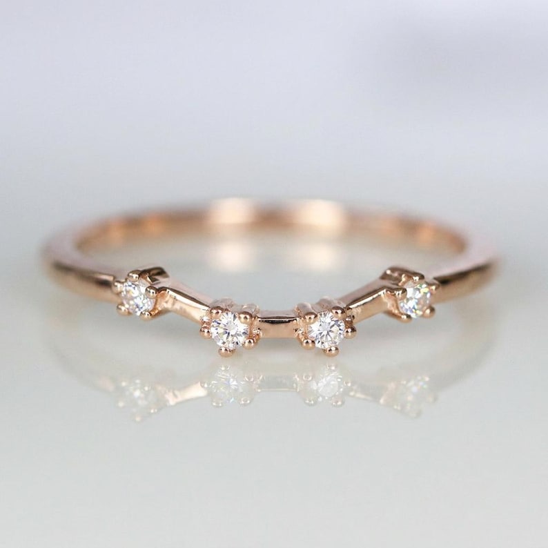 Puede incluir: Anillo de banda de oro rosa con cinco peque&ntilde;as piedras preciosas claras. El anillo tiene un dise&ntilde;o curvo con las piedras preciosas dispuestas en una fila en la parte superior. El anillo se muestra sobre una superficie reflectante.