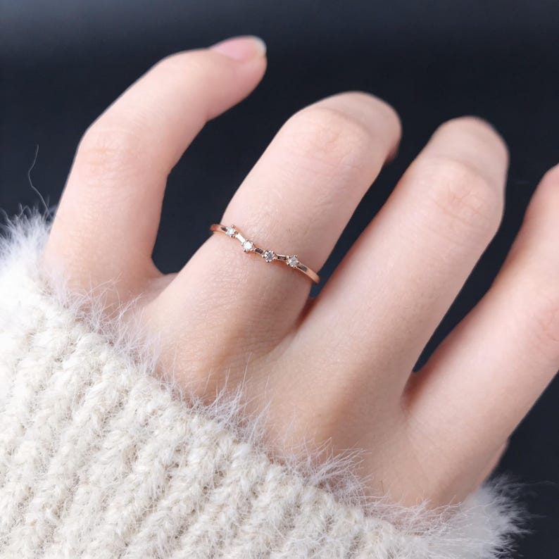 Puede incluir: Un delicado anillo de oro rosa con un dise&ntilde;o curvo. El anillo presenta una serie de peque&ntilde;as gemas brillantes engastadas a lo largo de la curva. El anillo se muestra en el dedo de una persona, destacando su estilo elegante y minimalista.
