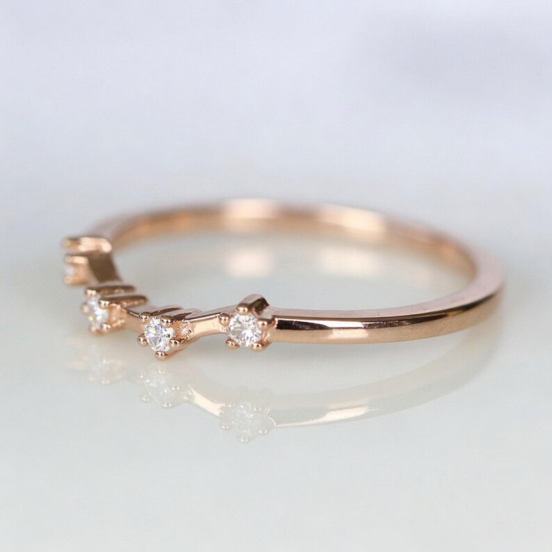 Puede incluir: Anillo de banda de oro rosa con tres peque&ntilde;as piedras preciosas transparentes. El anillo tiene un dise&ntilde;o delicado y curvado. Las piedras preciosas est&aacute;n espaciadas uniformemente a lo largo de la parte superior de la banda.