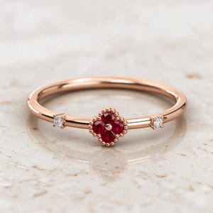 May include: A rose gold ring featuring a central four-petaled flower motif set with four small red rubies. The band is accented on either side of the floral centerpiece by a single small, clear, round-cut white diamond set in a rose gold bezel.