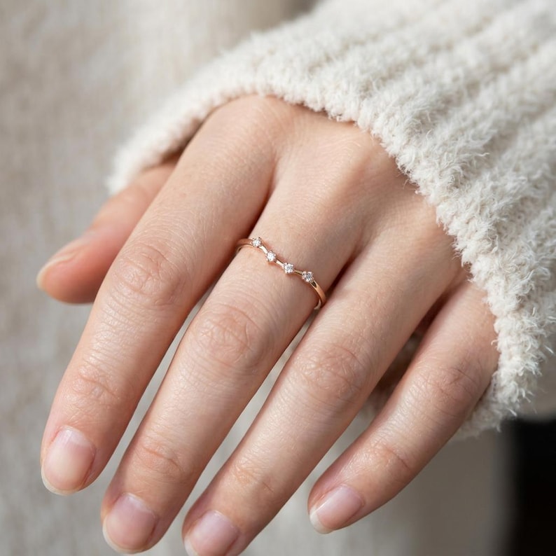 Puede incluir: Un delicado anillo de oro con cinco peque&ntilde;as piedras preciosas transparentes engastadas en un dise&ntilde;o curvo. El anillo se lleva en un dedo y la mano descansa sobre un su&eacute;ter blanco.
