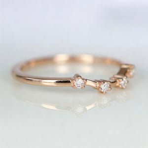 Puede incluir: Anillo de oro rosa con un dise&ntilde;o delicado. El anillo presenta peque&ntilde;os diamantes brillantes engastados en un patr&oacute;n de estrella a lo largo de la banda. El anillo se muestra sobre una superficie reflectante, destacando su acabado pulido.