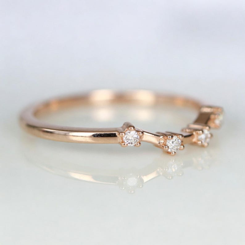 Puede incluir: Anillo de oro rosa con un dise&ntilde;o delicado. El anillo presenta peque&ntilde;os diamantes brillantes engastados en un patr&oacute;n de estrella a lo largo de la banda. El anillo se muestra sobre una superficie reflectante, destacando su acabado pulido.