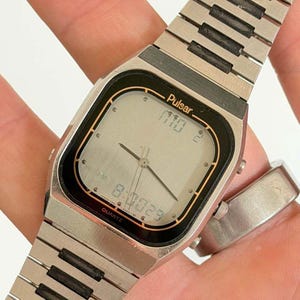 Può includere: Un orologio da polso al quarzo analogico-digitale Pulsar color argento con quadrante quadrato in metallo spazzolato, lunetta nera e lancette centrali. Il display digitale mostra l'ora 8:00:29. Ha un cinturino a maglie in metallo argentato.