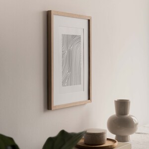 K&ouml;nnte beinhalten: Ein gerahmter Schwarzwei&szlig;-Linienkunst-Druck mit abstrakten Formen h&auml;ngt an einer wei&szlig;en Wand. Eine beige Keramikvase und ein runder beigefarbener Beh&auml;lter stehen auf einem wei&szlig;en Tisch mit einer gr&uuml;nen Pflanze im Vordergrund.